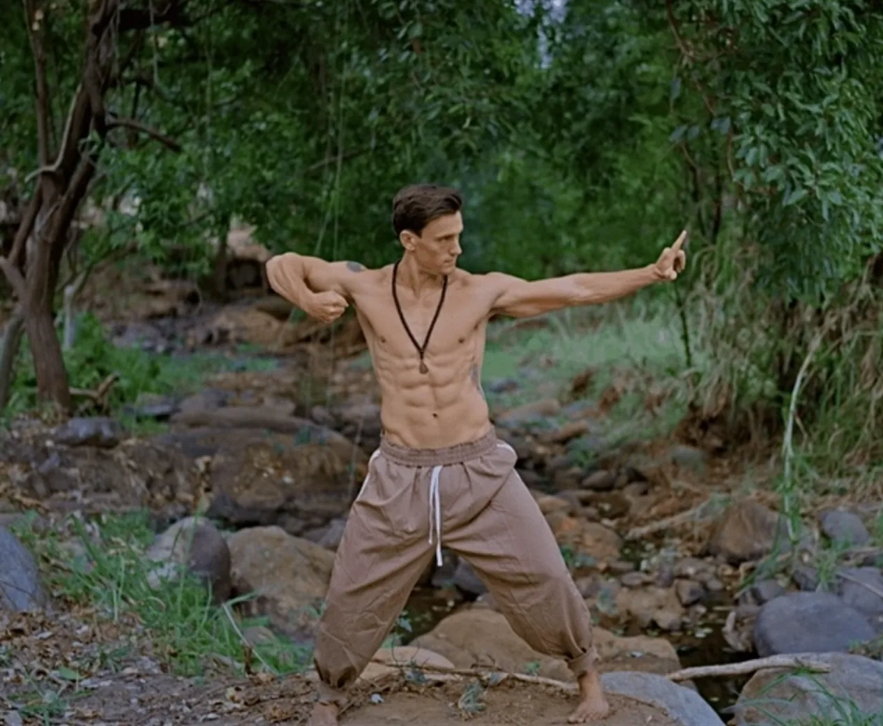 The coach barefoot on river rocks, holding a Qi Gong posture with one arm extended
